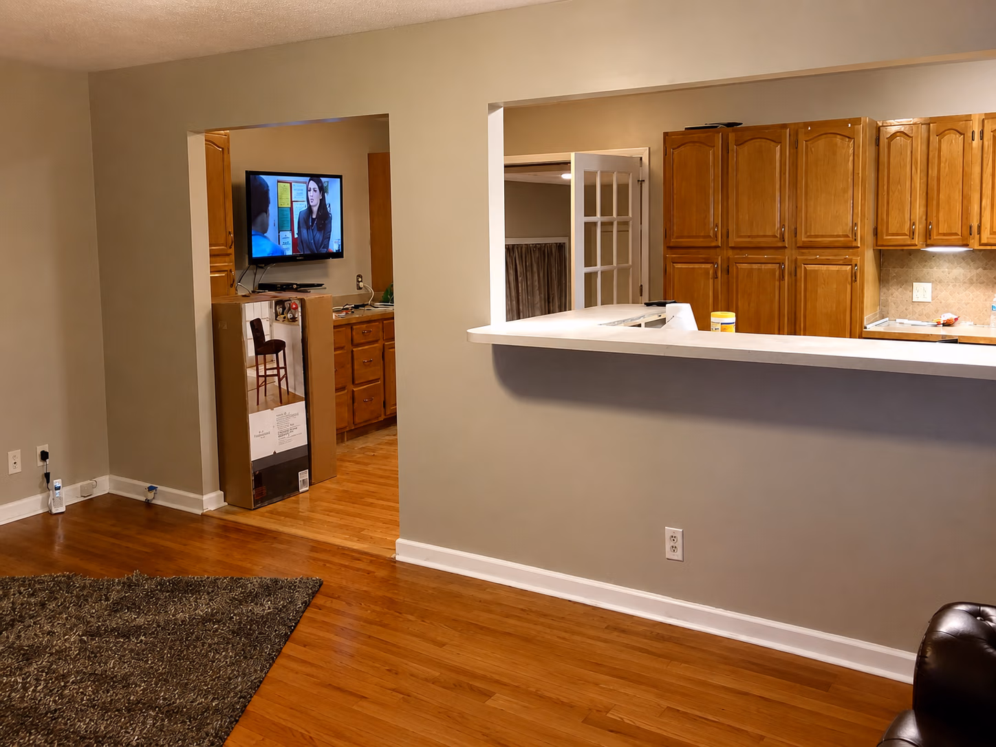 Open living space with hardwood floors, kitchen with wooden cabinets, and TV visible through doorway.