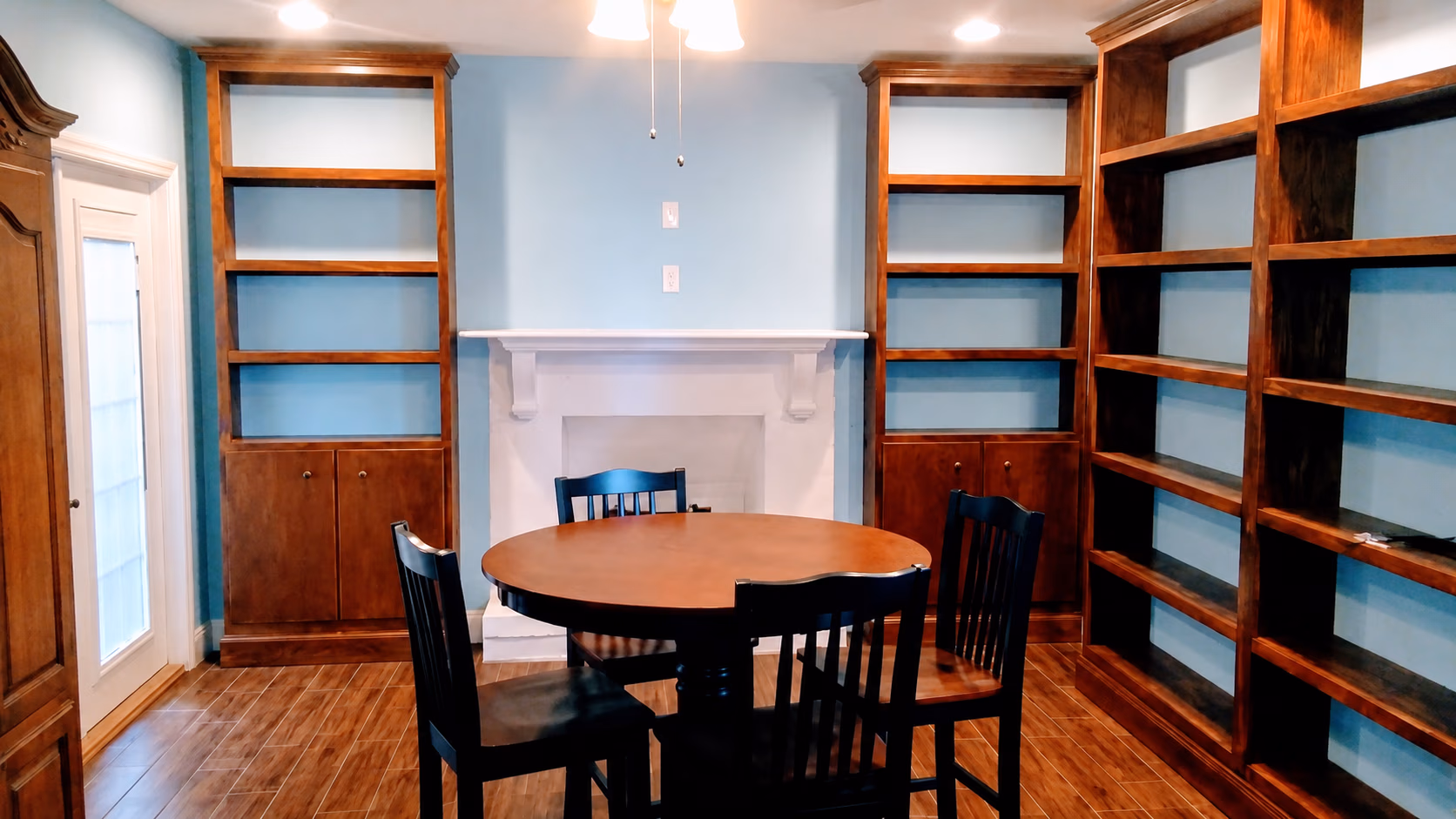 Room with wooden round table, four black chairs, built-in empty wooden shelves, and white fireplace.