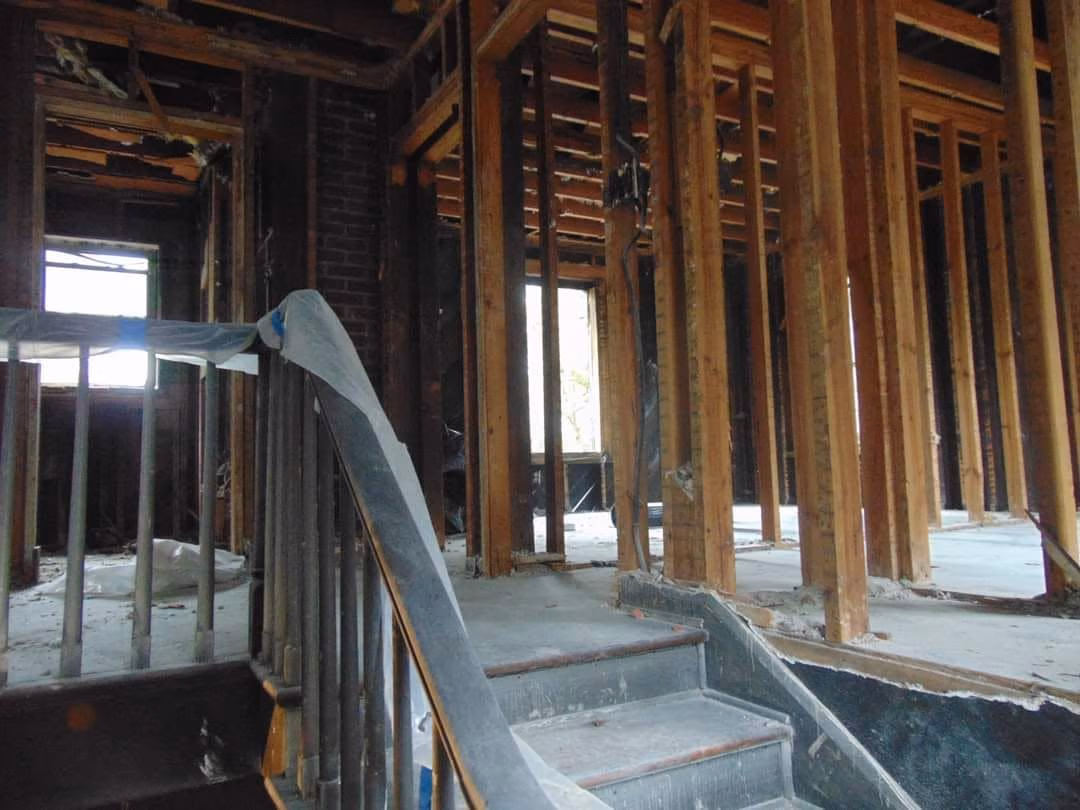 Interior of a house under renovation showing exposed wooden framing and a staircase wrapped with protective material.