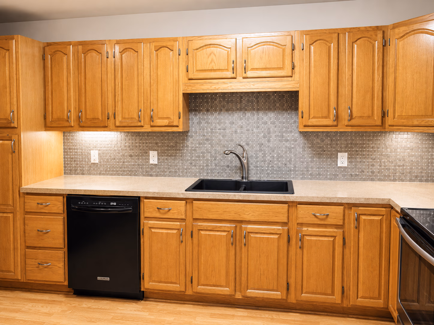 Kitchen with oak cabinets, black dishwasher, black sink, beige countertop, and tiled backsplash.