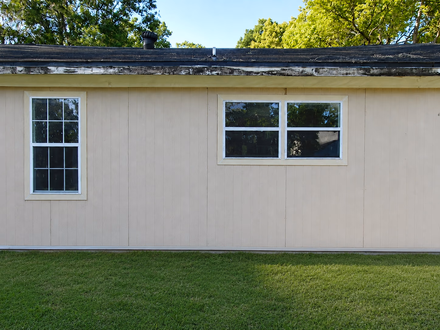 Beige exterior wall with two white-framed windows and green lawn in front under trees.