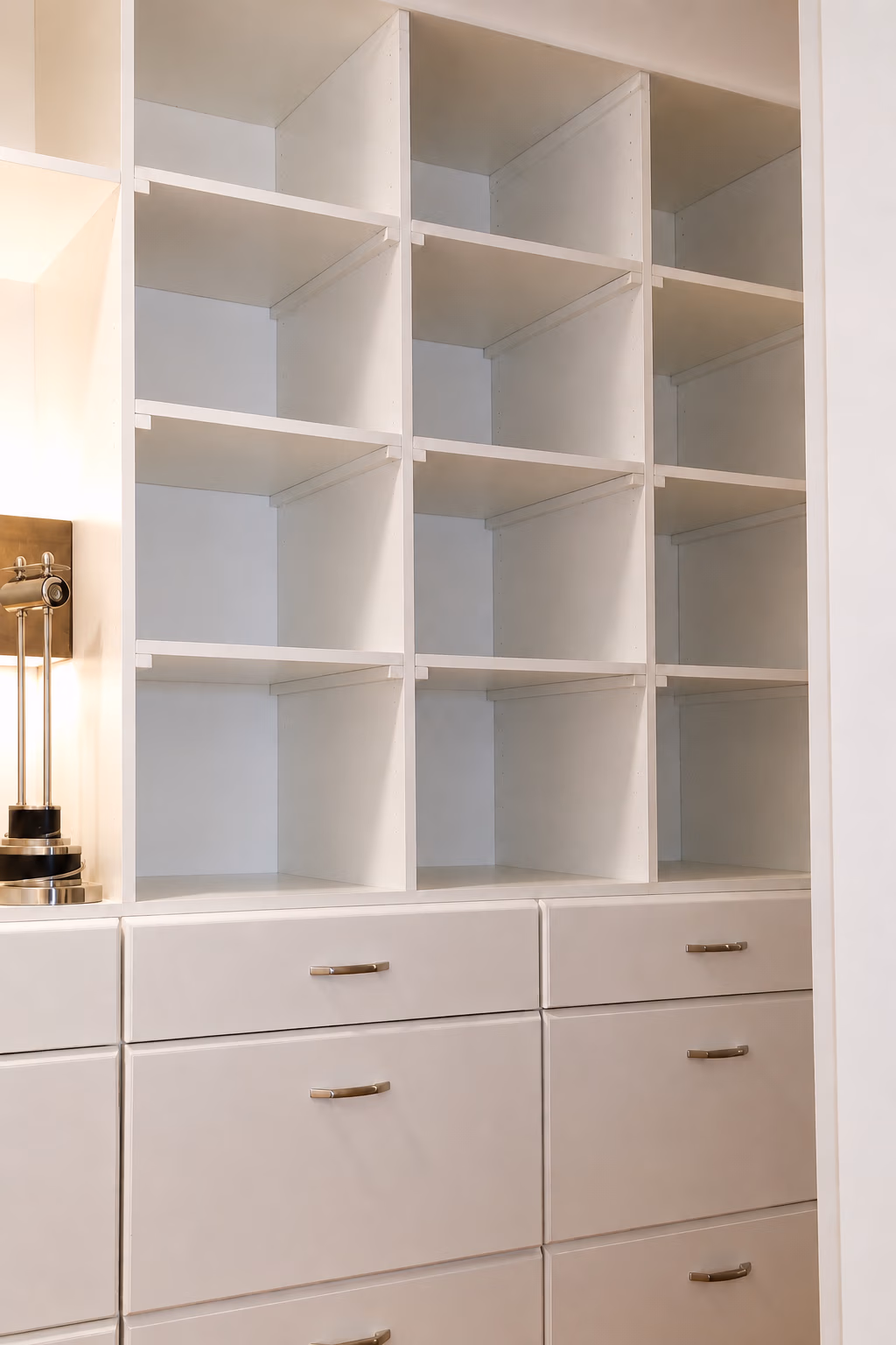 Empty white shelving unit with drawers beneath, next to a modern lamp on the left side.