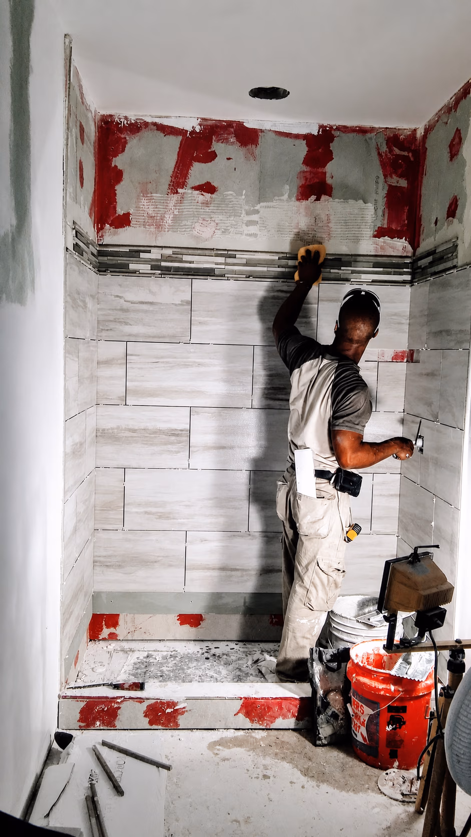 Worker installing beige tiles on shower walls with tools and supplies around him.