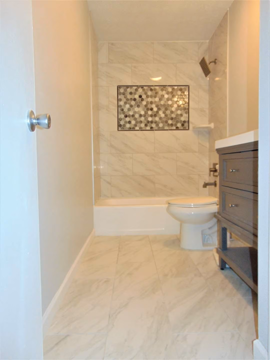 Modern bathroom with marble tile floor, white bathtub, toilet, dark vanity, and mosaic tile shower wall.