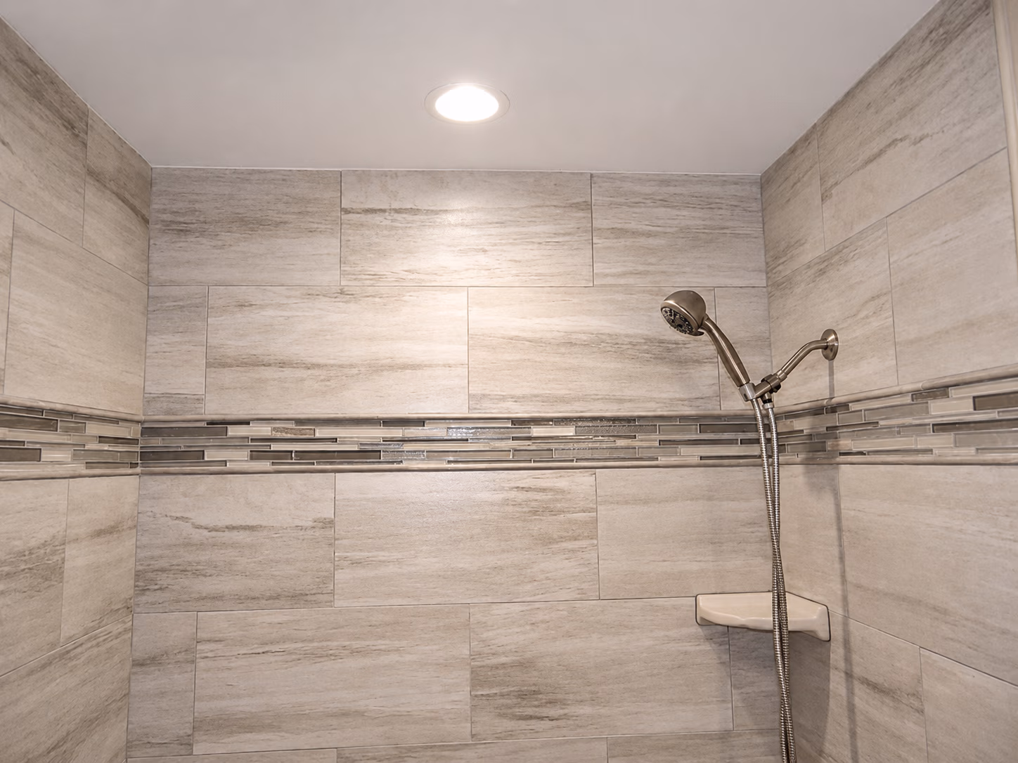 Shower area with beige rectangular tiles and a horizontal band of glass mosaic tiles with a mounted showerhead.