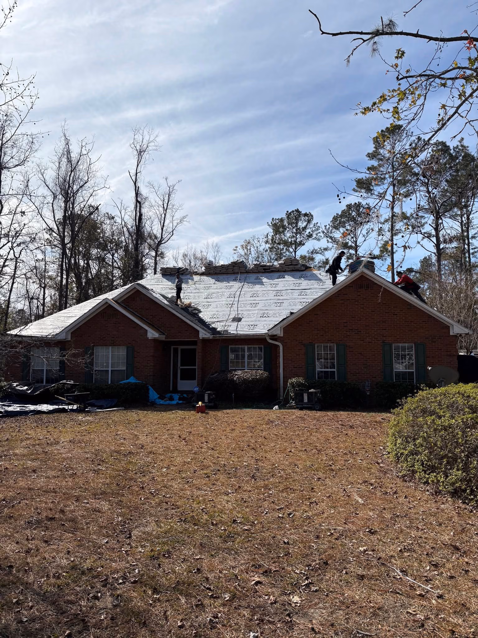Workers installing roofing materials on a brick house under a partly cloudy sky surrounded by trees.