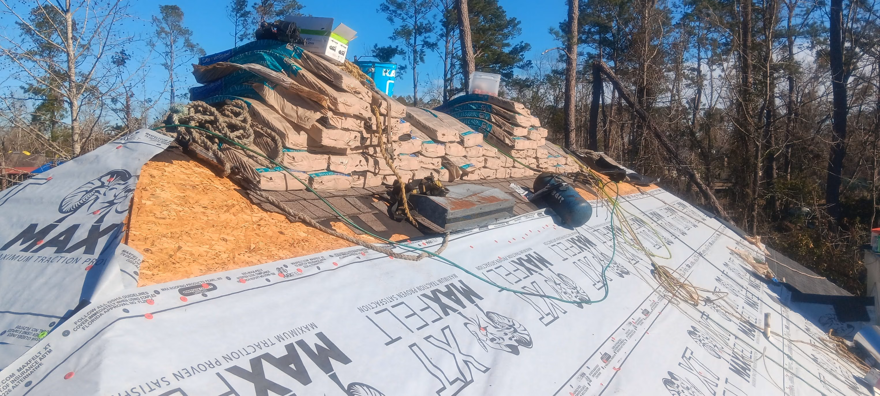 Roof under construction with stacked shingles, roofing felt, wood panels, and tools in a wooded area.