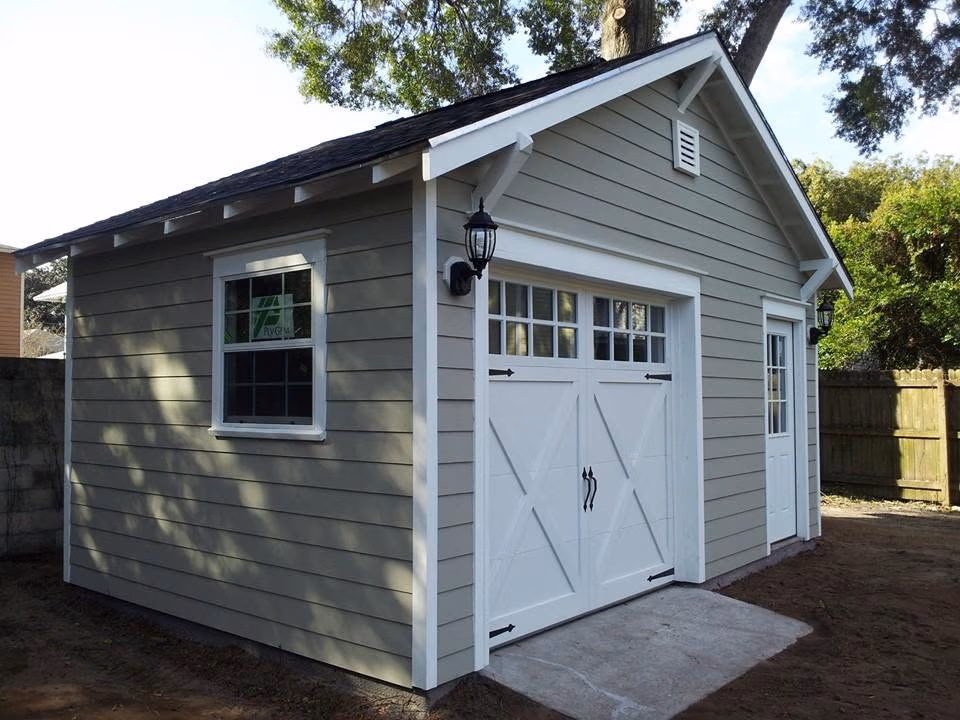 Small gray shed with white trim, double garage door, side window, and entry door under trees.