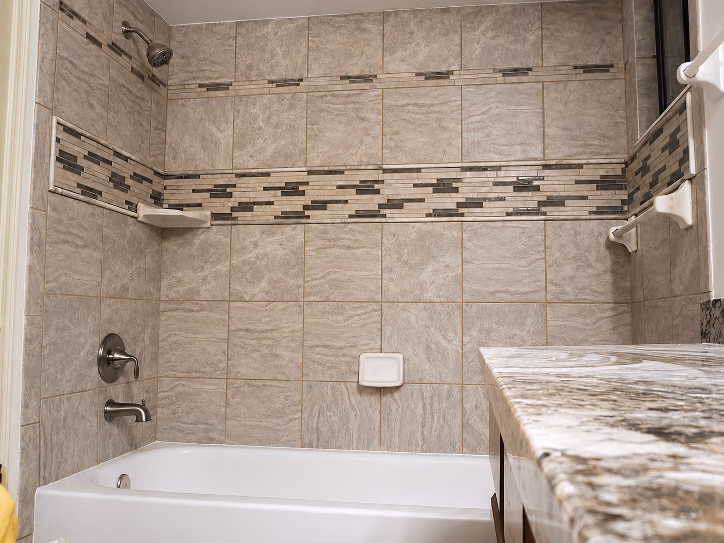 Bathroom with beige tiled shower and bathtub, granite countertop, chrome faucet, and soap holders.