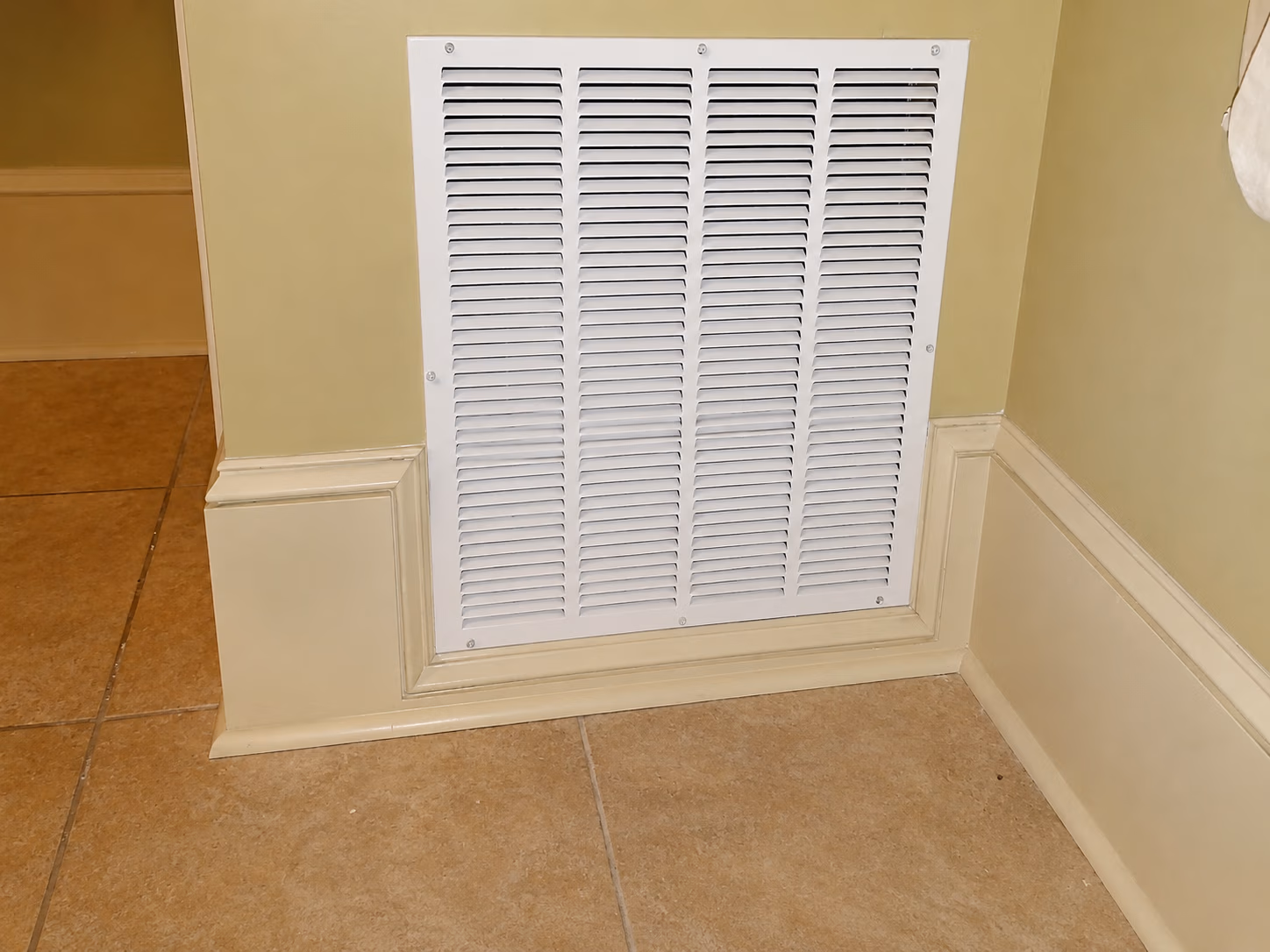 White wall vent grille with horizontal slats on a beige wall above tiled floor baseboards.