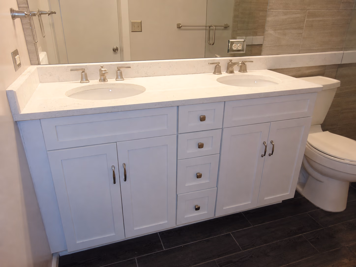 Bathroom vanity with double sinks, white cabinets, silver faucets, and a toilet on dark tile floor.