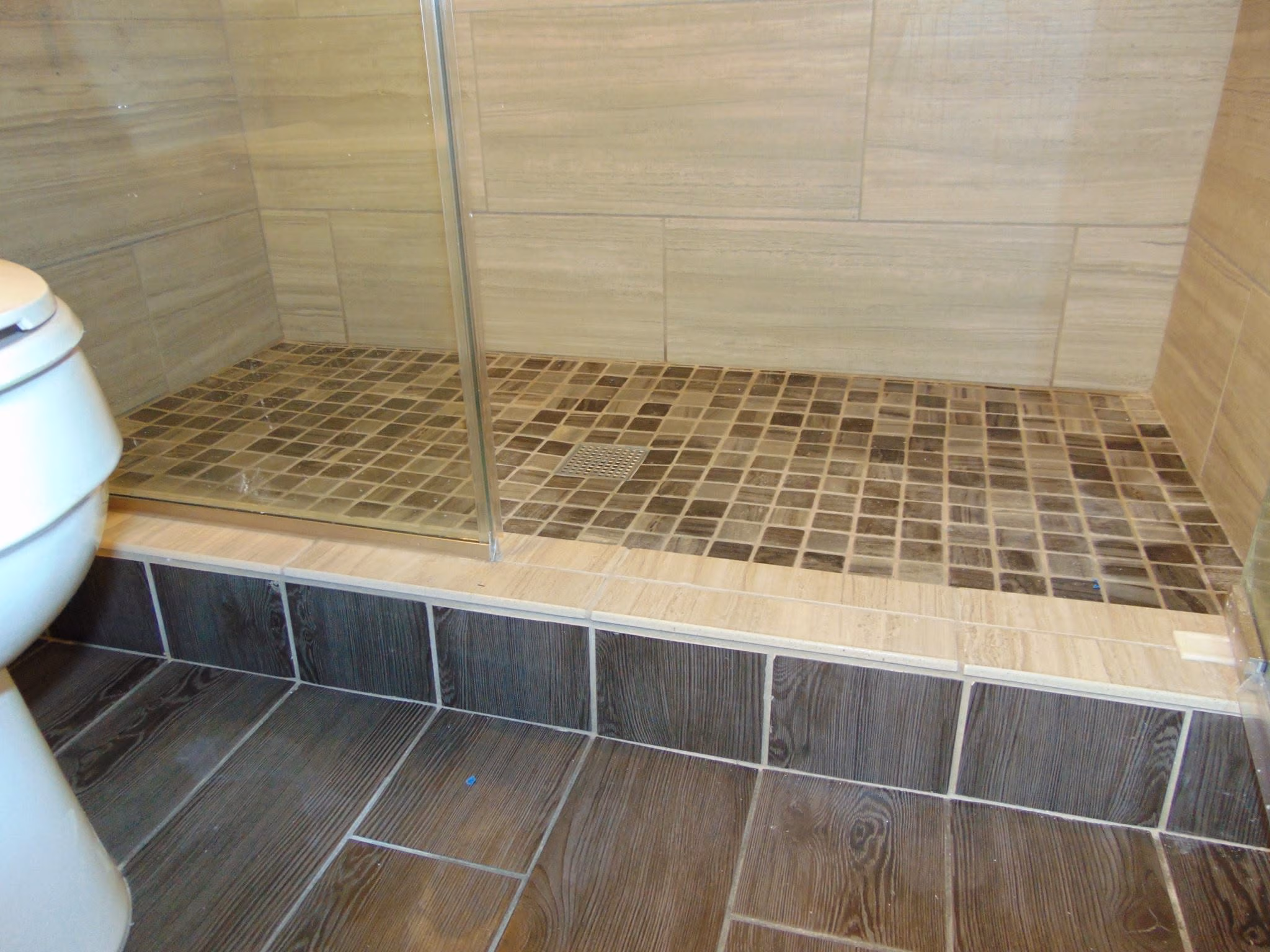 Bathroom floor with wood-look tiles, a raised shower area with small square tiles and glass sliding door.