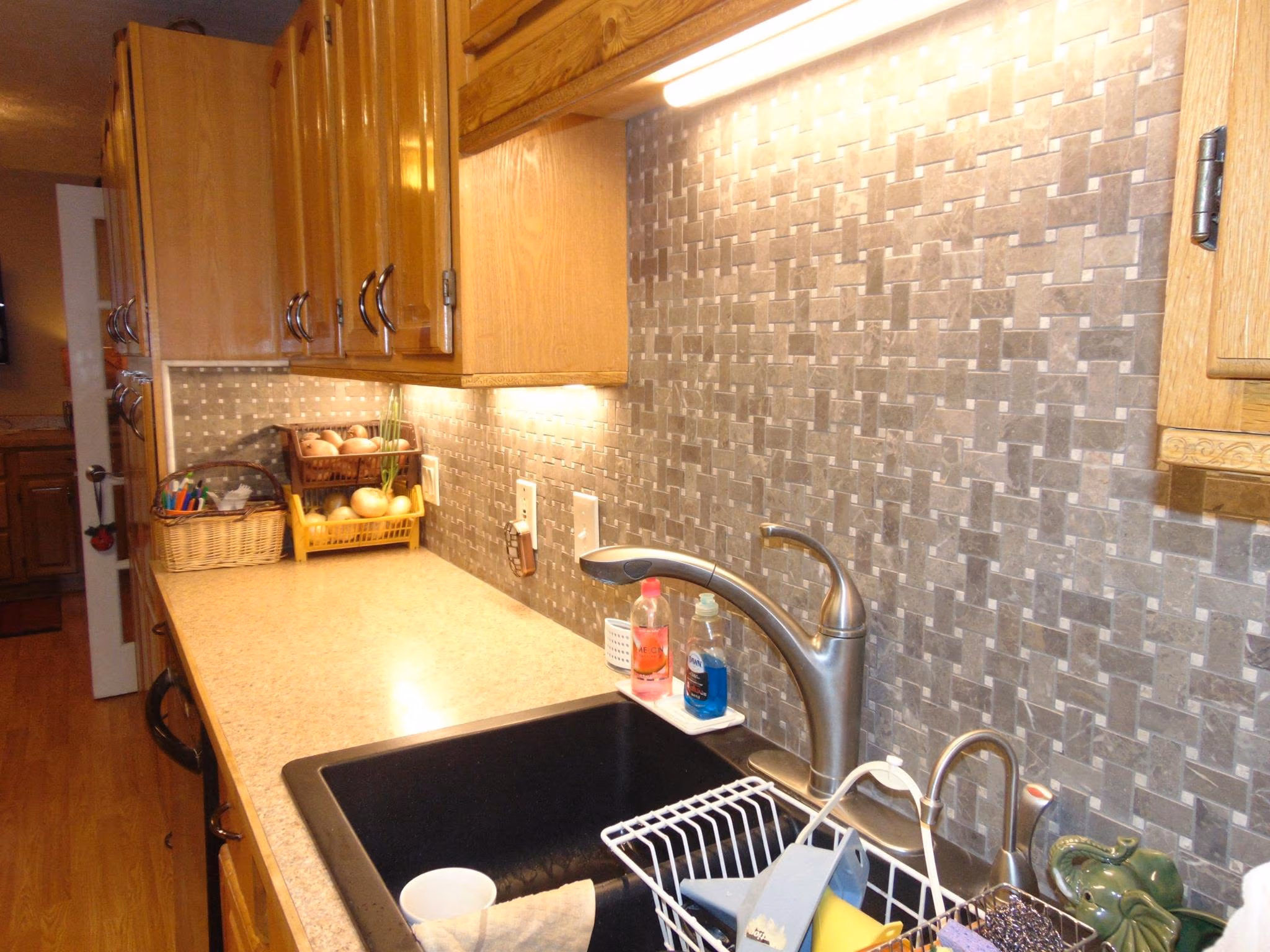Kitchen counter with wooden cabinets, a sink, faucet, tiled backsplash, and baskets with onions and utensils.