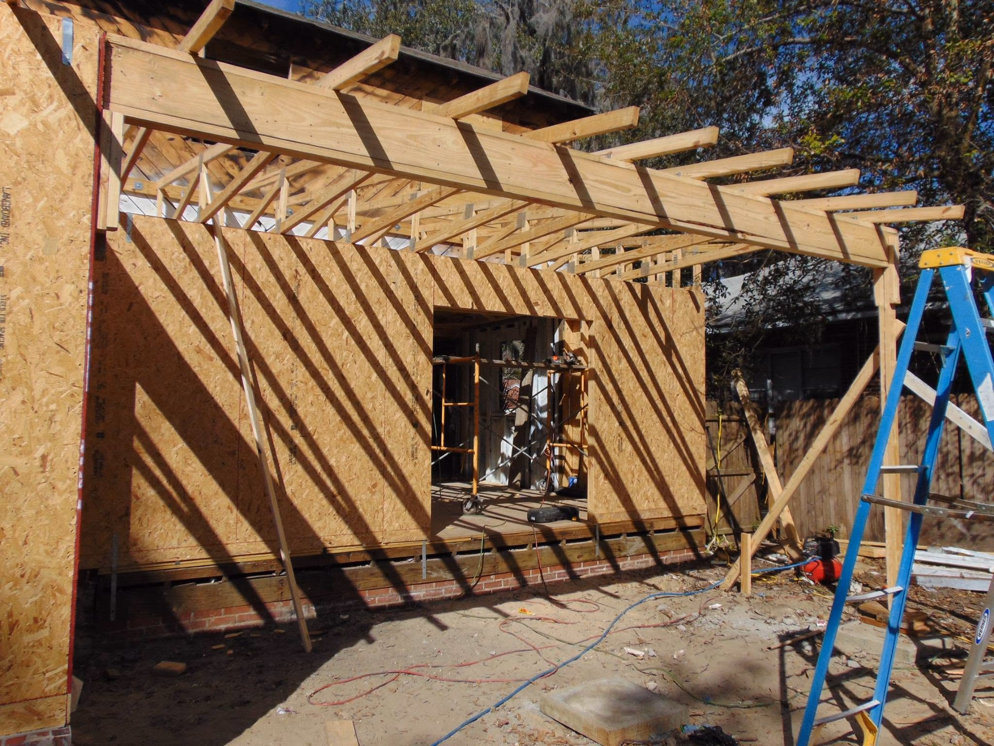 Construction site showing wooden framing and OSB sheathing with shadows and a blue ladder on dirt ground.