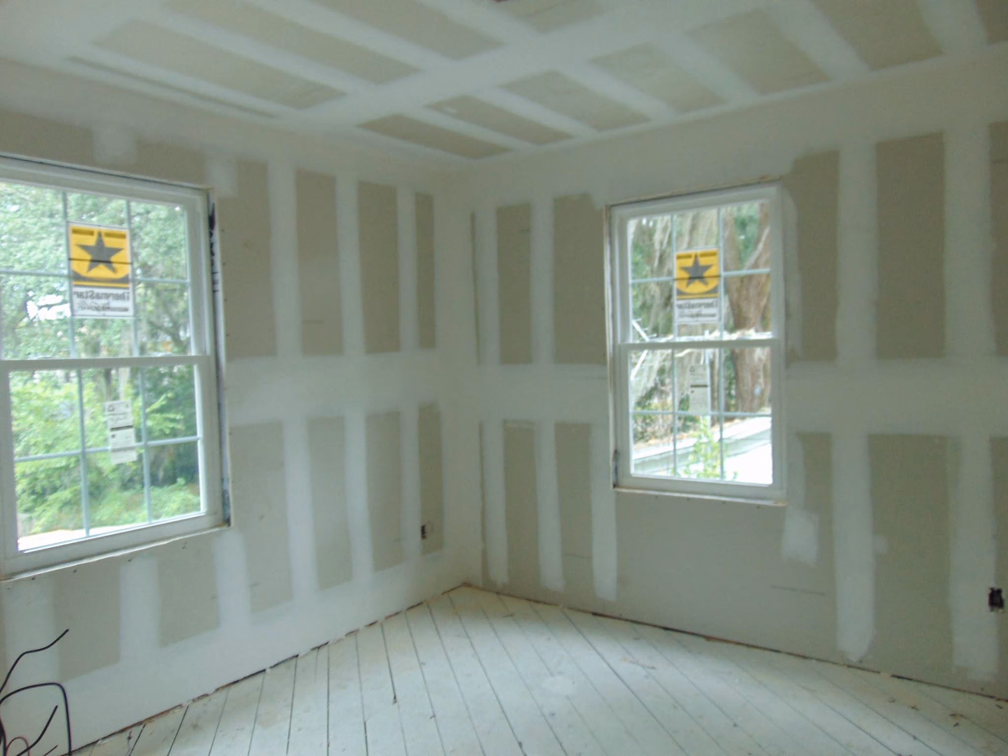Empty room with unfinished drywall walls and two windows showing trees outside.