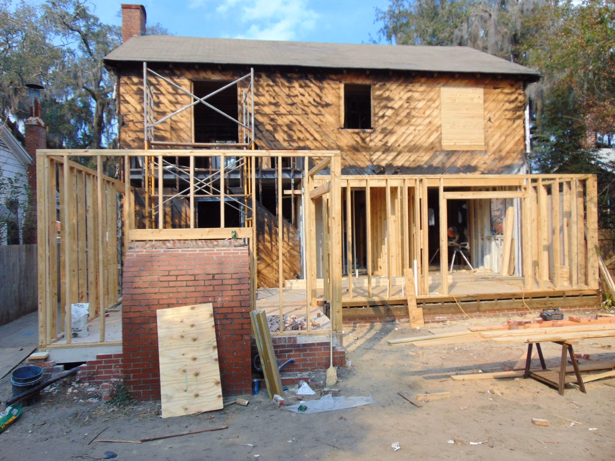House under construction with wooden frame and brick chimney in front on a dirt lot.