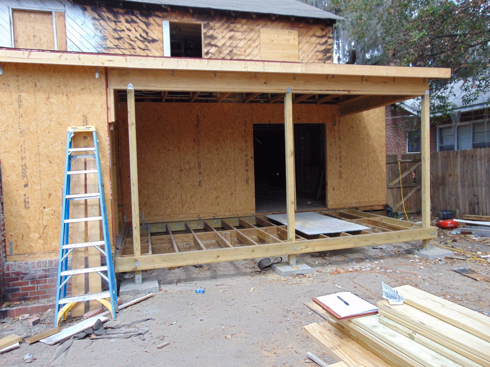Wooden frame structure under construction with ladder, plywood panels, and construction materials nearby.