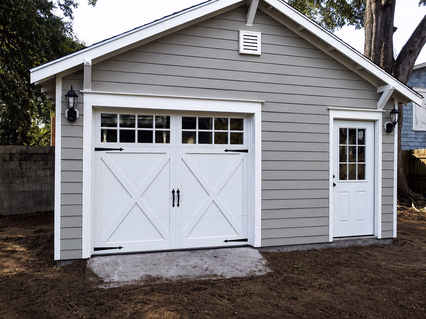 Gray building with white double garage door and adjacent white entry door with glass panes.