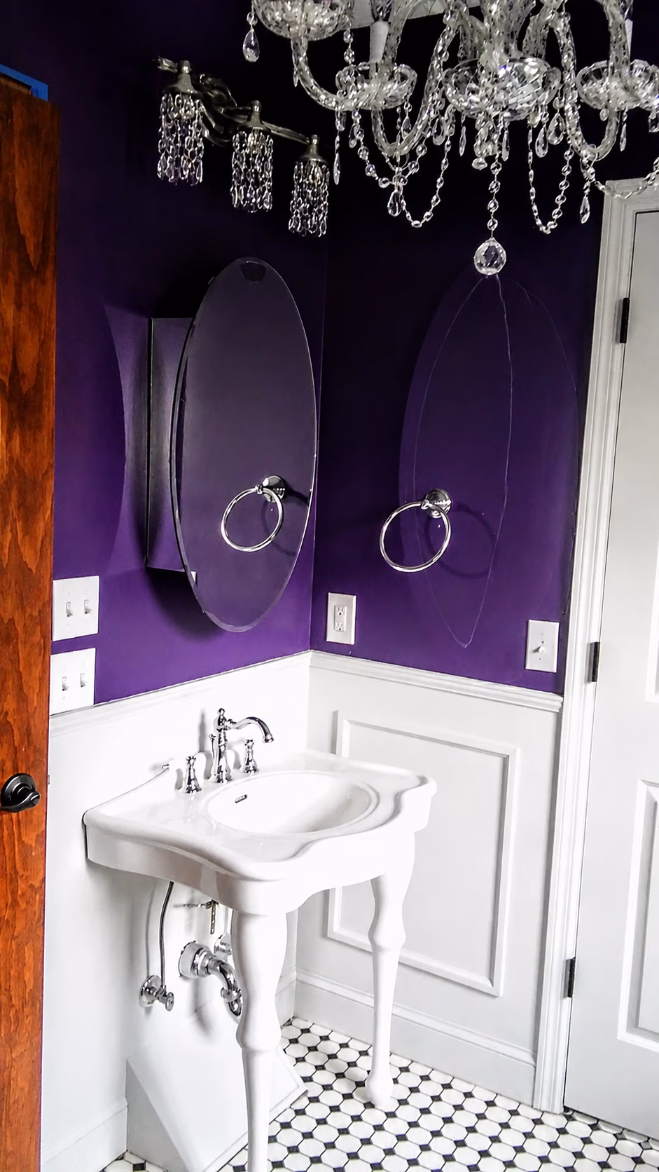 Bathroom corner with white pedestal sink, oval mirror, purple walls, and crystal chandelier.