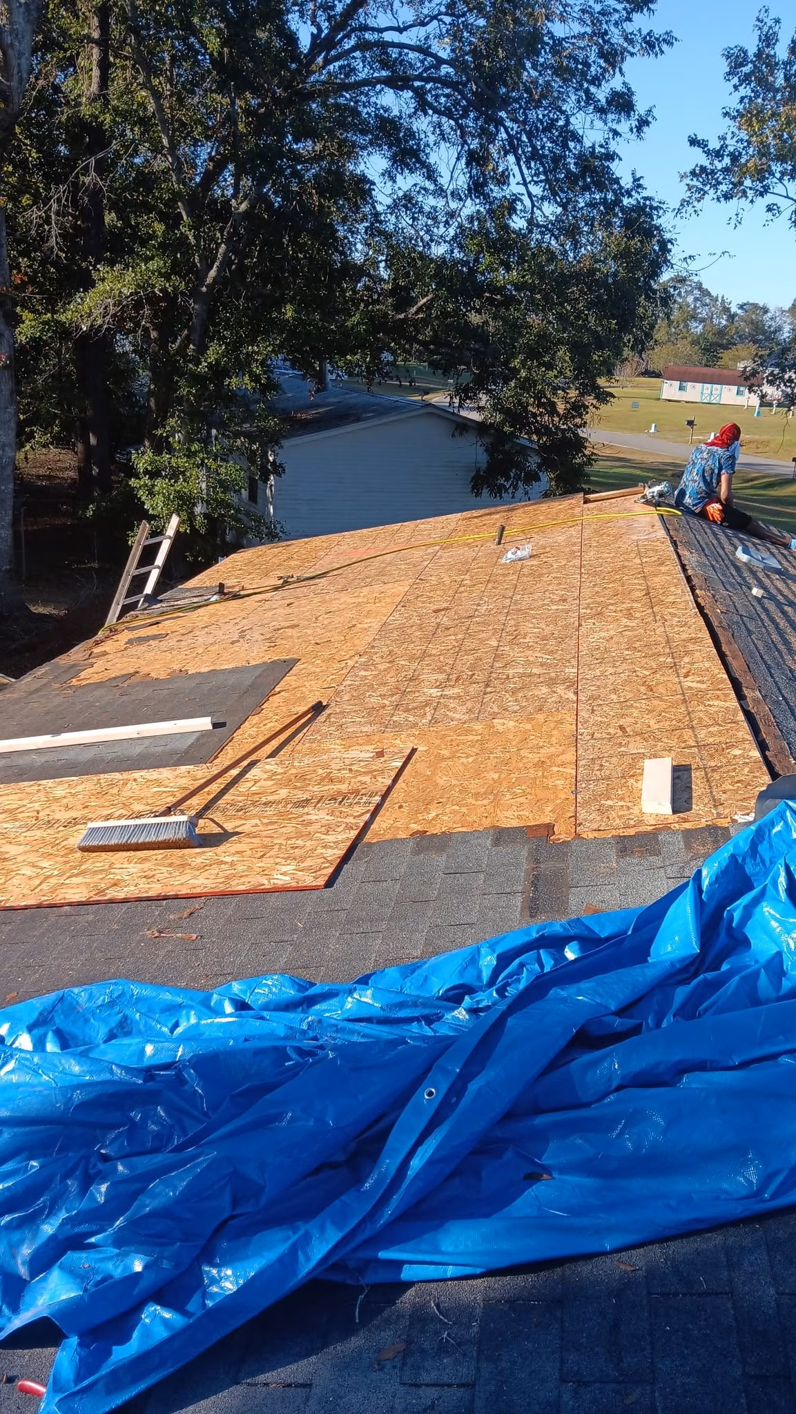 Roof under repair with plywood sheets, a person working, a broom, and a blue tarp covering part of the roof.