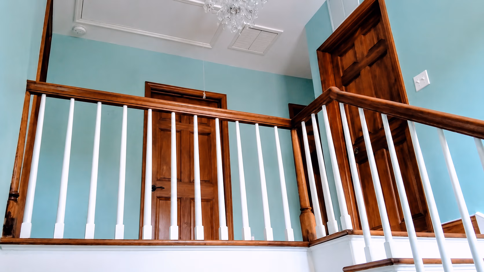 Indoor wooden railing with white spindles beside closed wooden doors on teal walls.