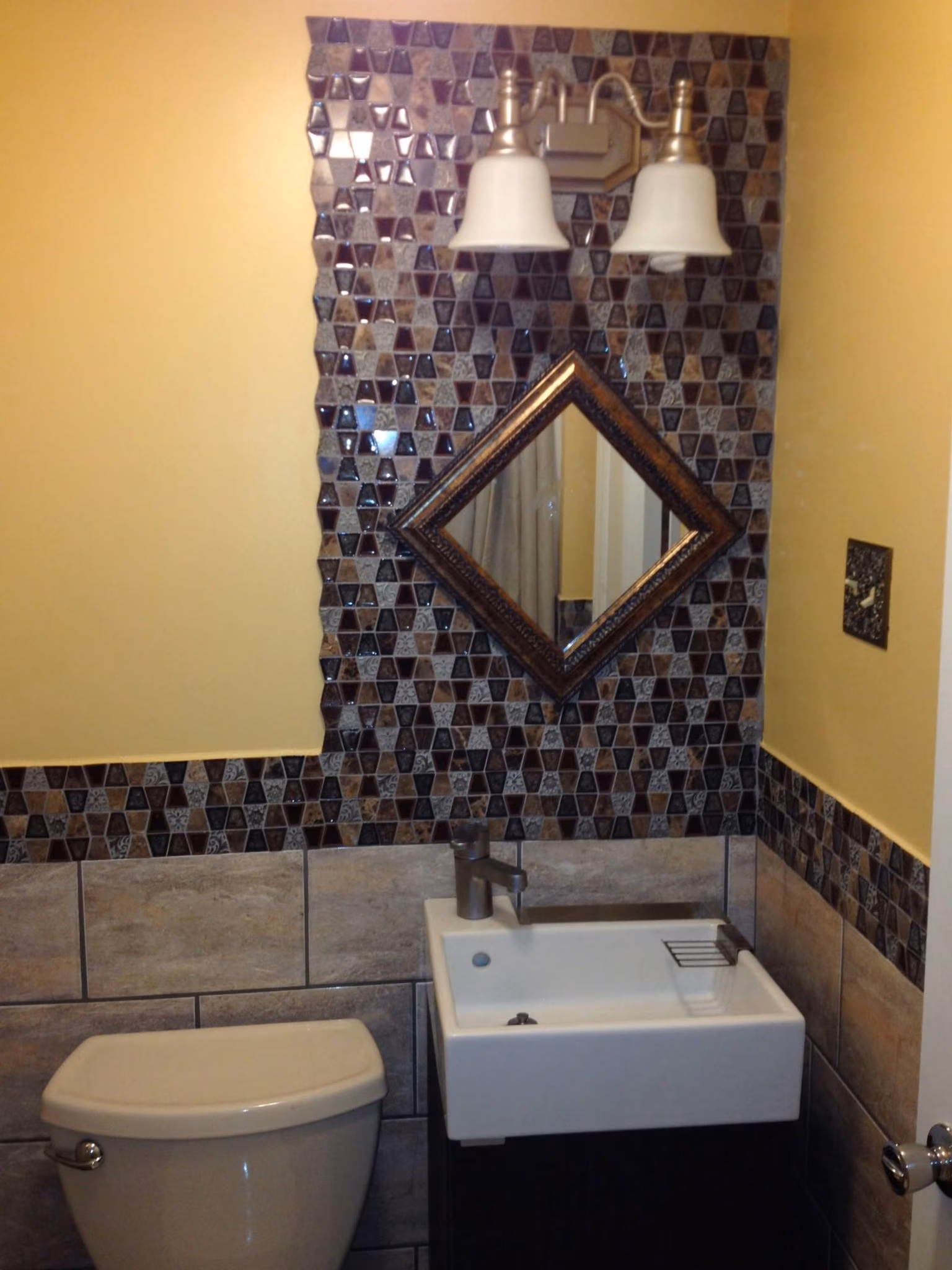 Small bathroom with beige toilet, white sink, diamond mirror, mosaic tile wall, and two light fixtures.