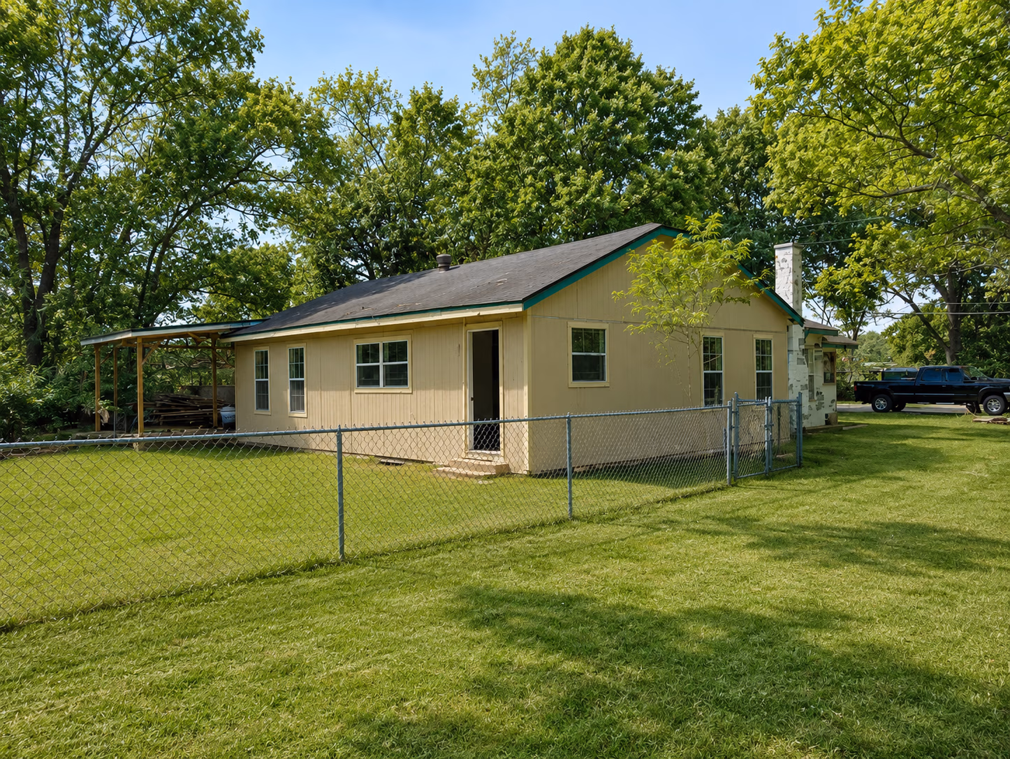 Single-story beige house with green trim surrounded by a chain-link fence and trees on a sunny day.
