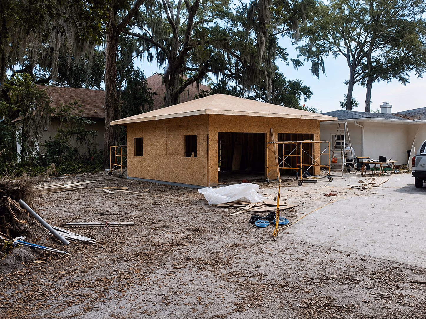 Wooden garage under construction with plywood walls and roof next to a white house in a yard.