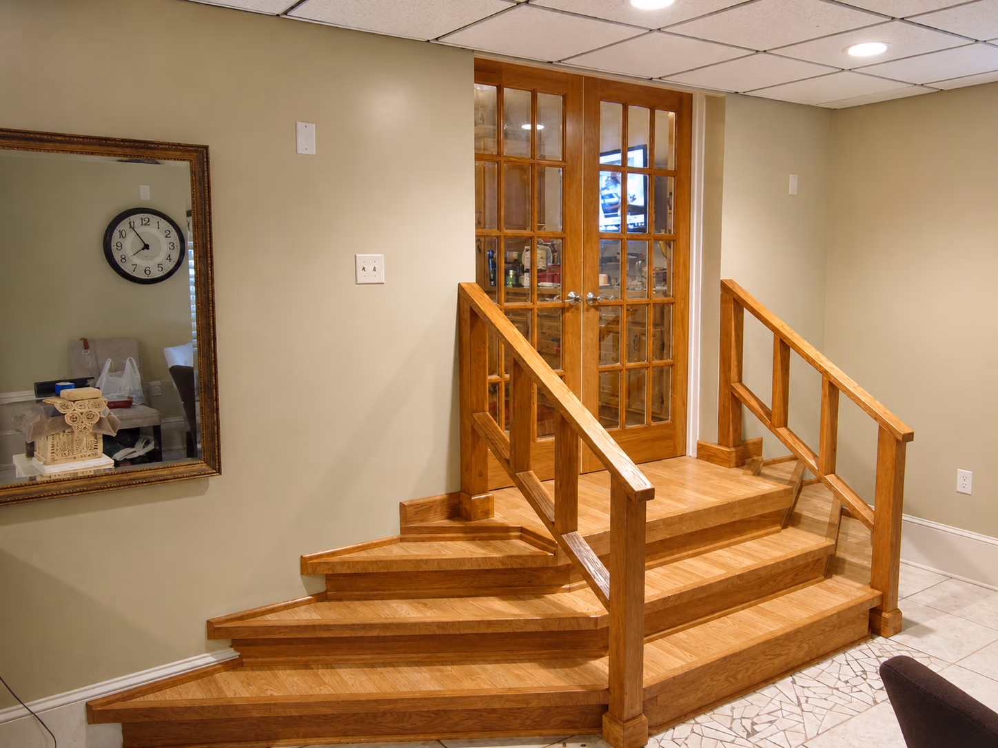 Wooden stairs with railings leading to glass double doors in a beige room with a large mirror and clock.