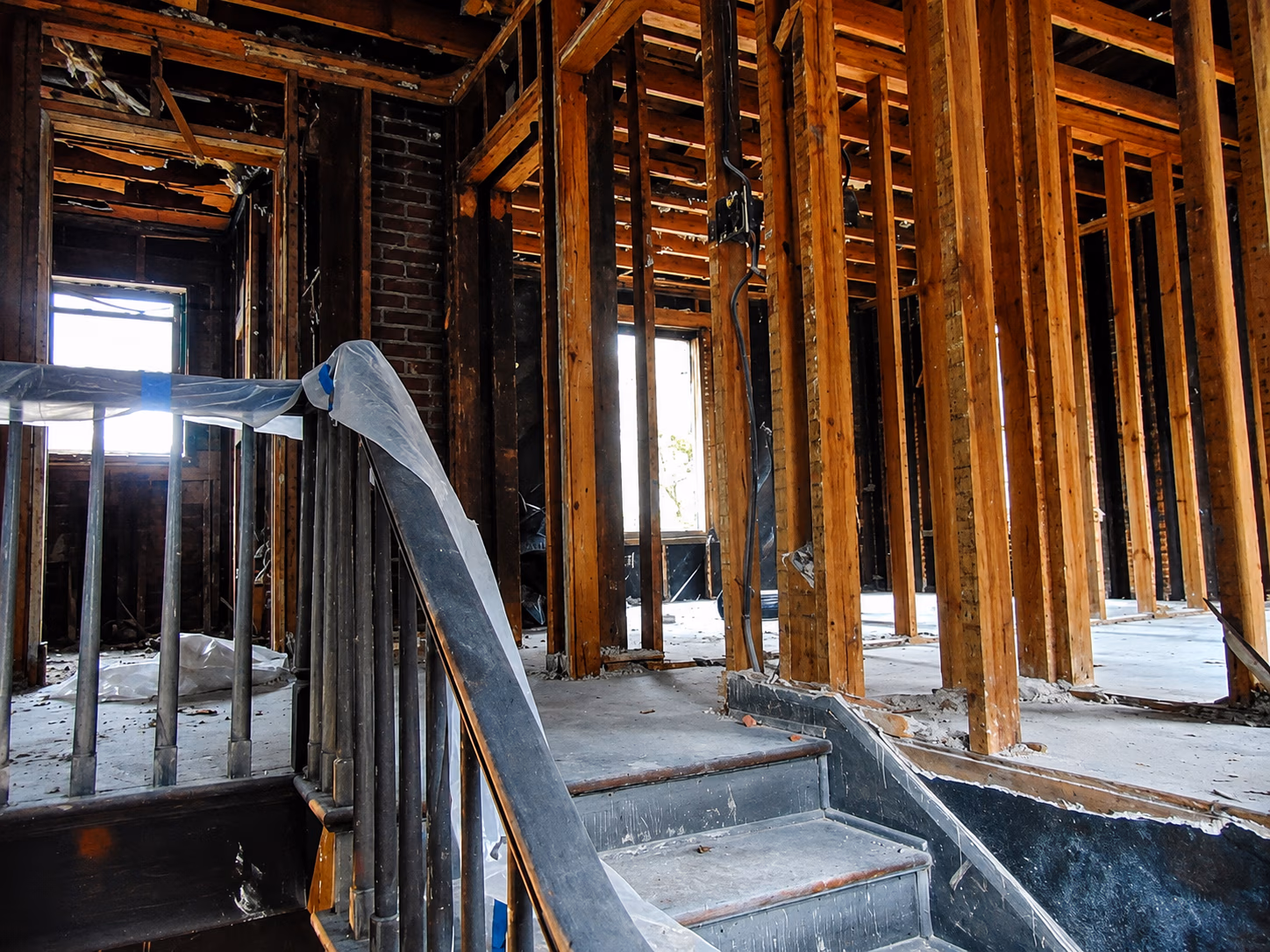 Interior of a house under renovation showing exposed wooden framing and a stairway with a plastic-covered handrail.