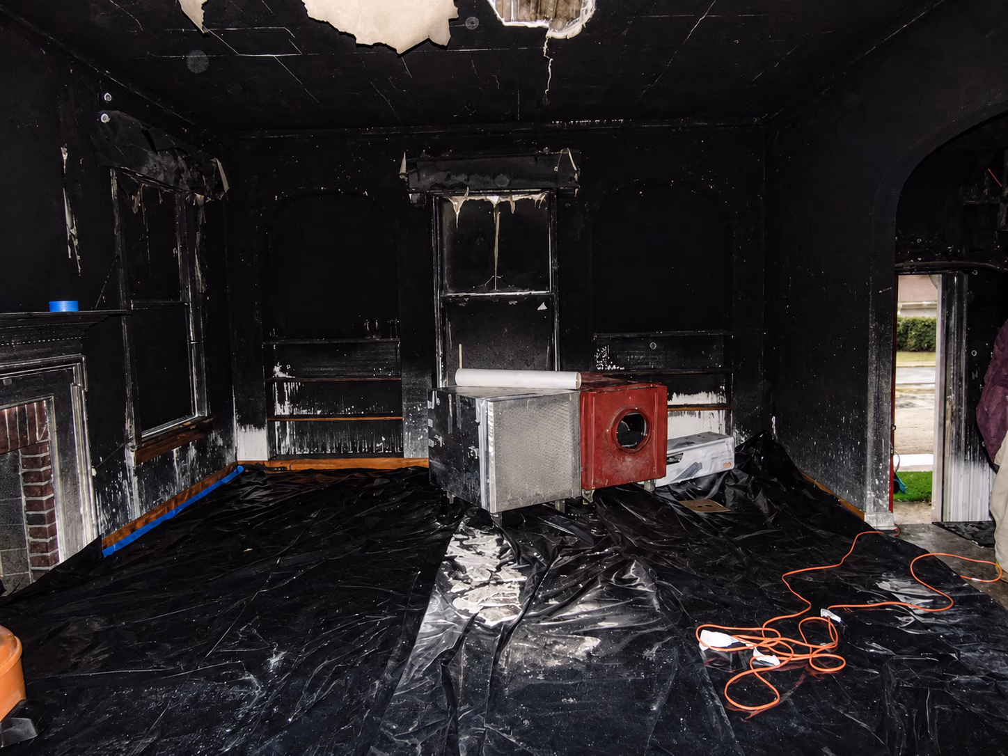 Room heavily damaged by fire, blackened walls and ceiling, covered floor with debris and equipment in the center.