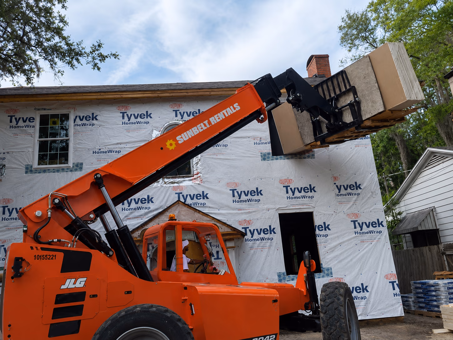 Orange Sunbelt Rentals forklift lifting plywood sheets at a house under construction with Tyvek wrap.