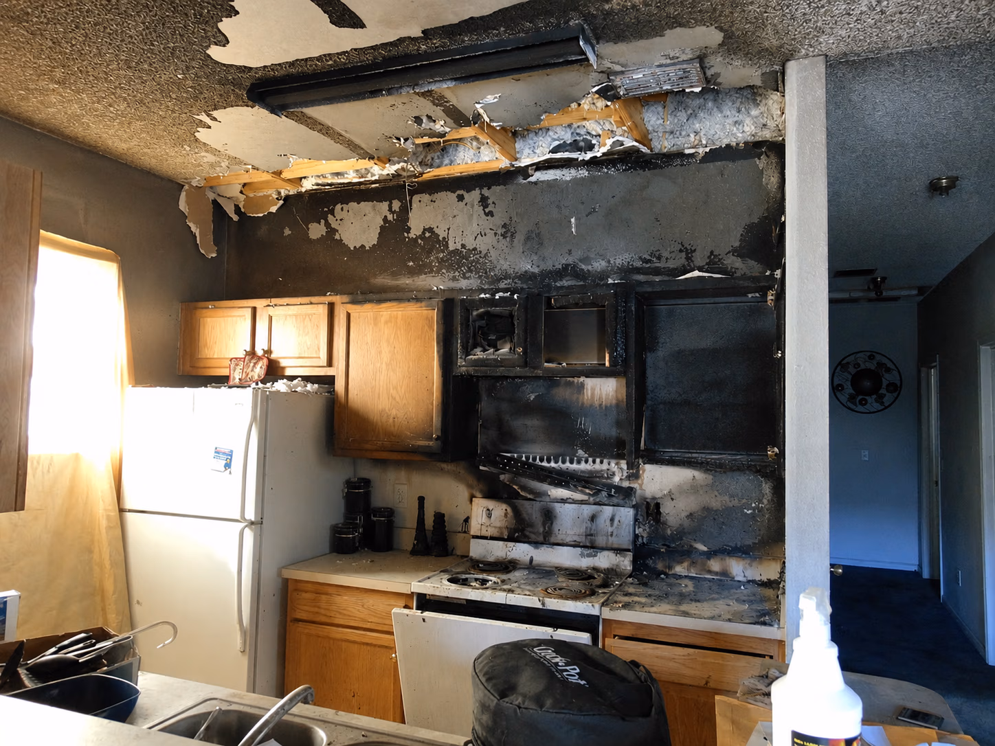 Kitchen with fire damage showing charred cabinets, stove, blackened walls, and peeled ceiling near window and refrigerator.