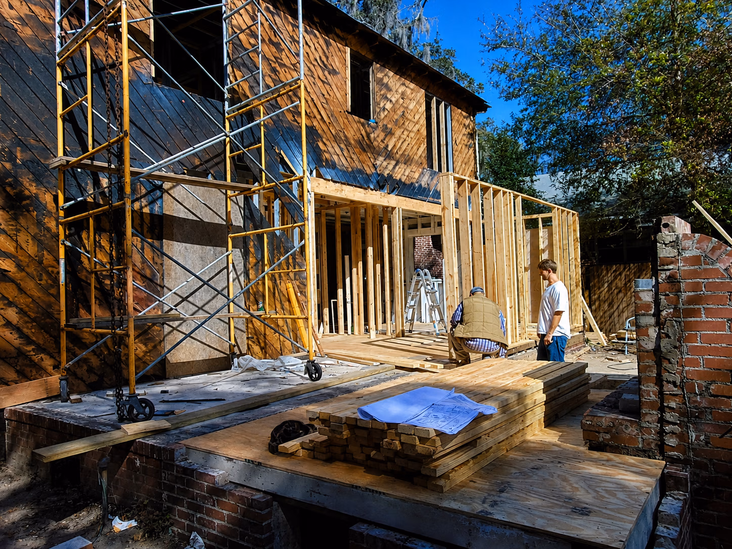 Two men working on wooden framework of house under construction with scaffolding and lumber.