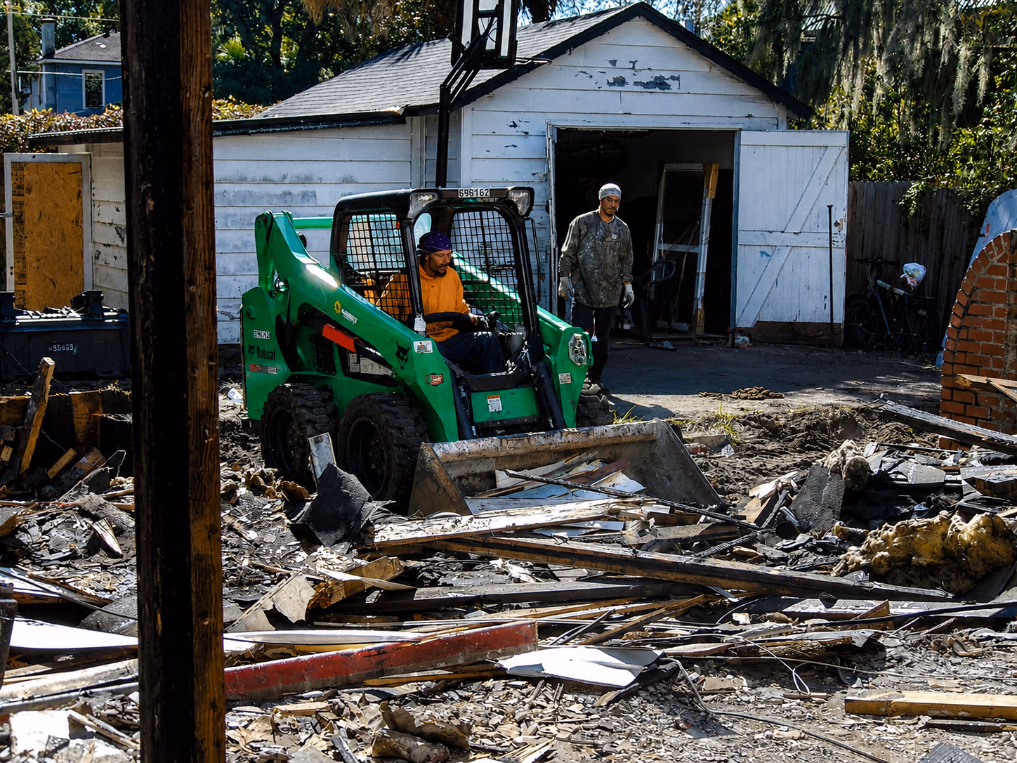 Worker operates a small green Bobcat skid-steer loader on a debris-filled construction site.