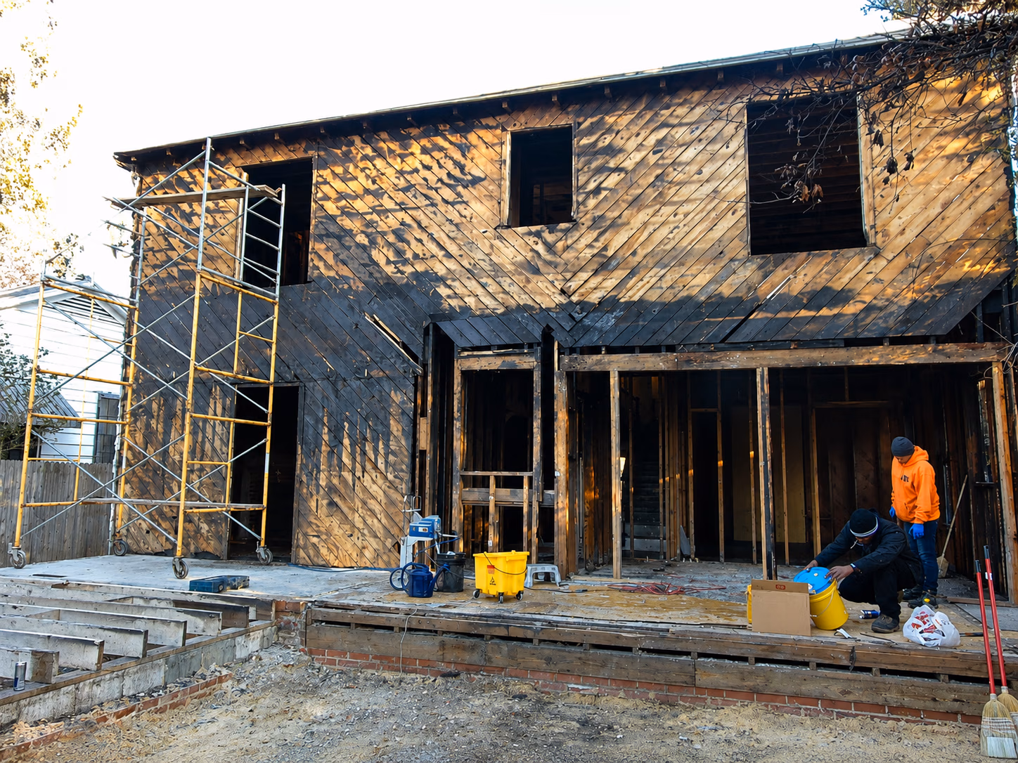 Two workers at a wooden house under renovation with scaffolding and construction tools outside.