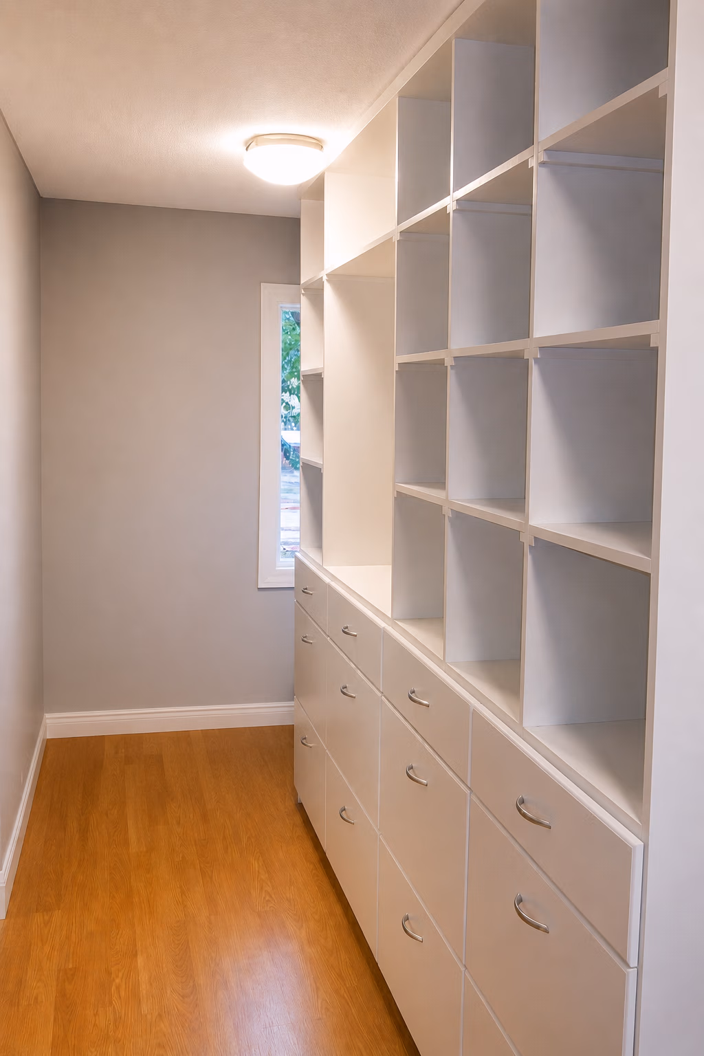 Empty white shelving units and cabinets along wood floor hallway with a window and ceiling light.