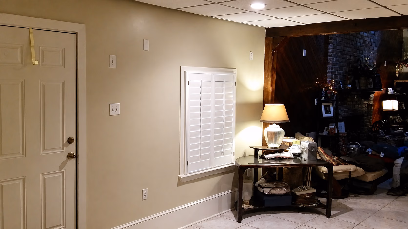 Room corner with beige walls, a closed door, white shuttered window, and lamp-lit wooden table with decor.