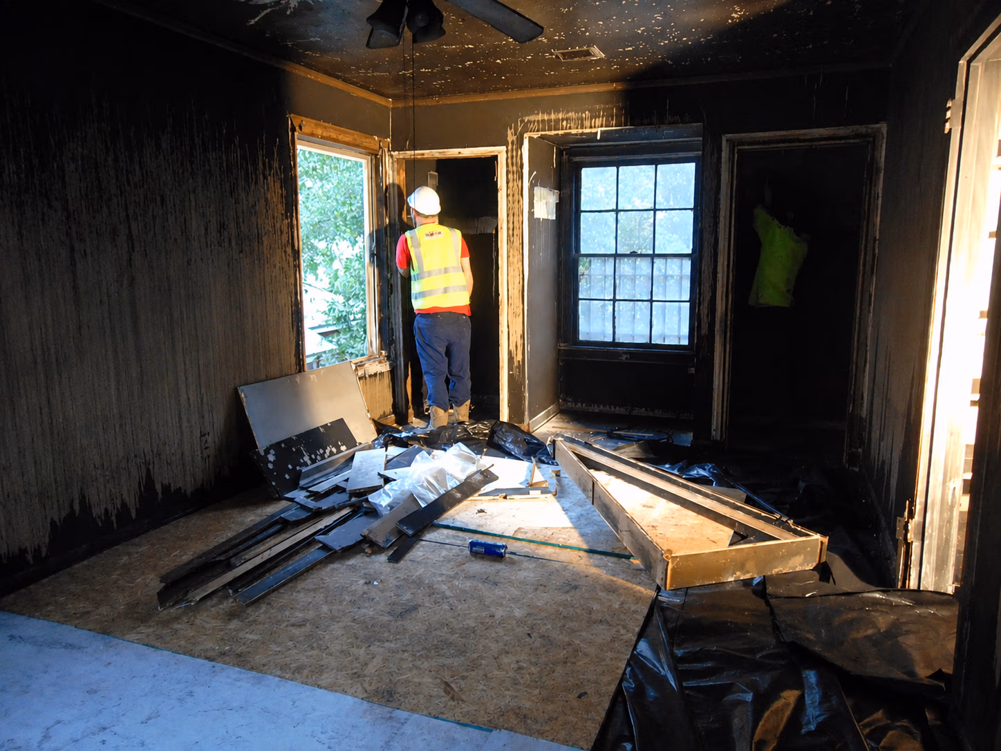 A construction worker inspects a fire-damaged room with charred walls and debris on the floor.