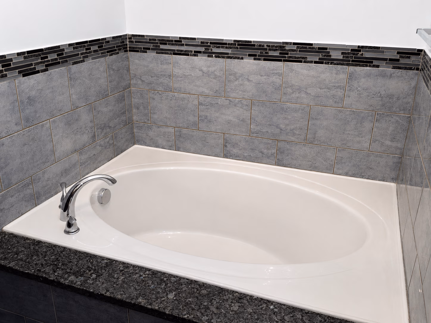 White built-in bathtub with chrome faucet surrounded by gray and black tiled walls and countertop.