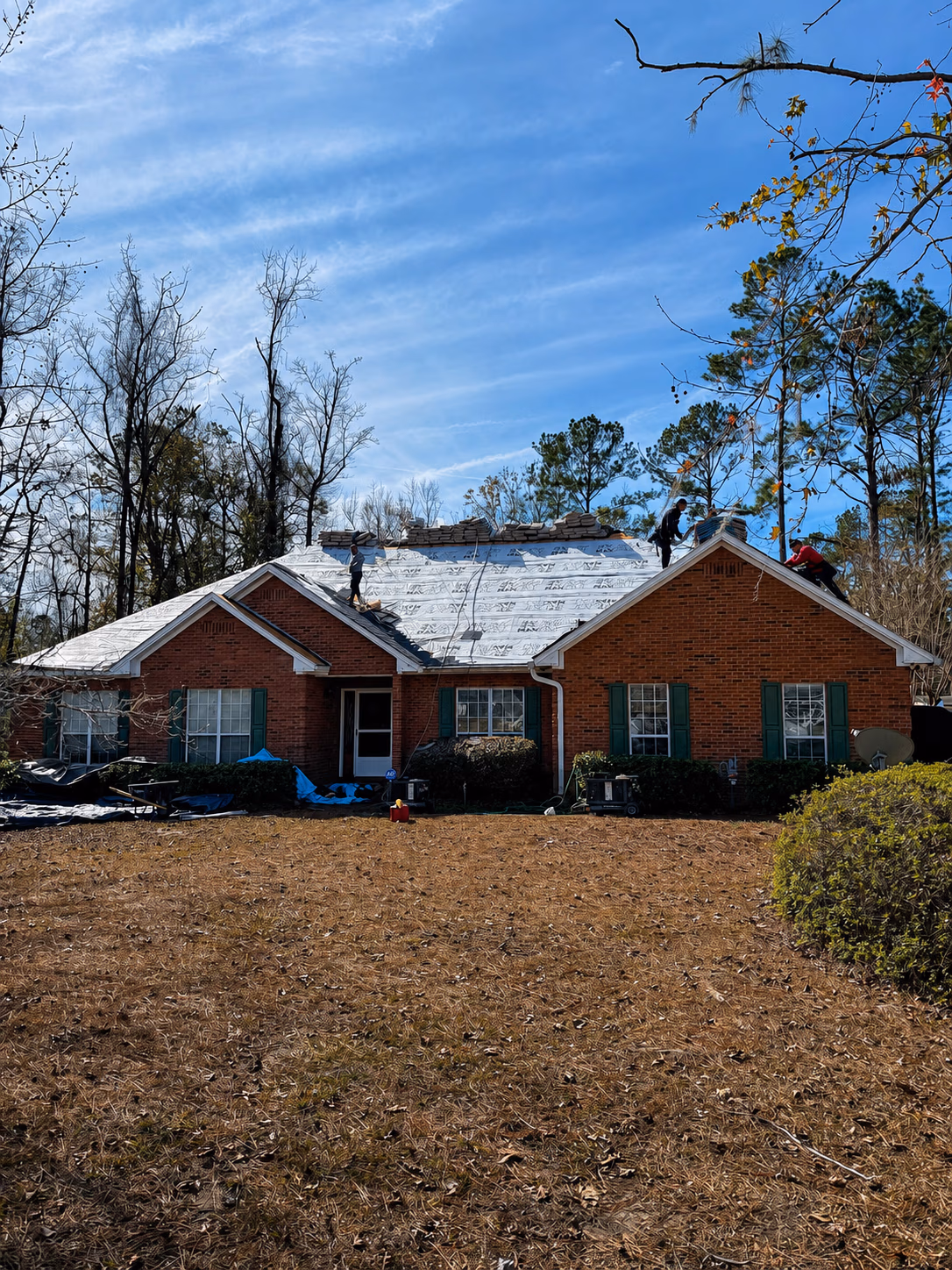 Three workers on the roof of a brick house installing underlayment on a clear day.