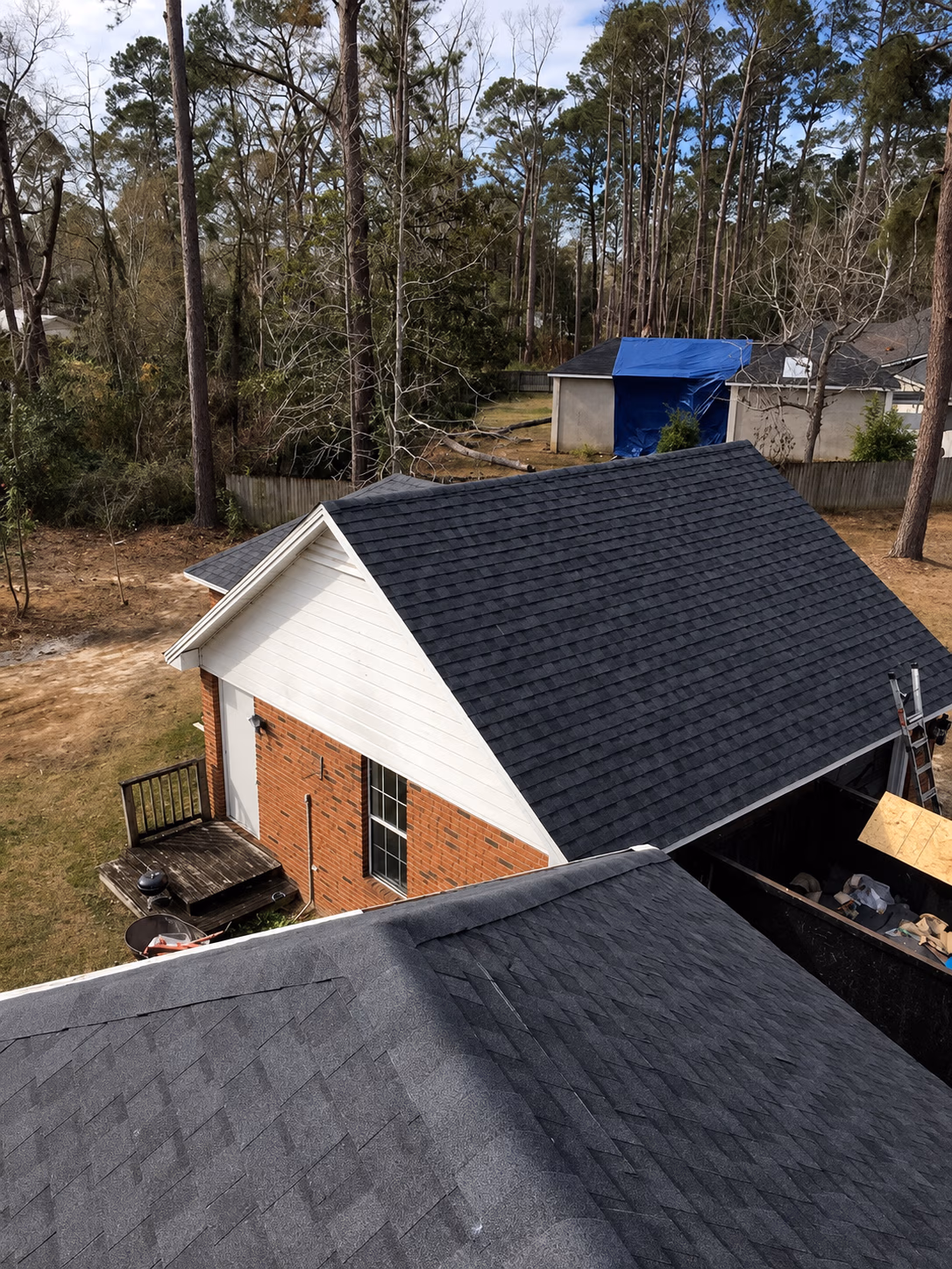 View of dark gray shingled roofs of residential buildings surrounded by tall trees and a yard.