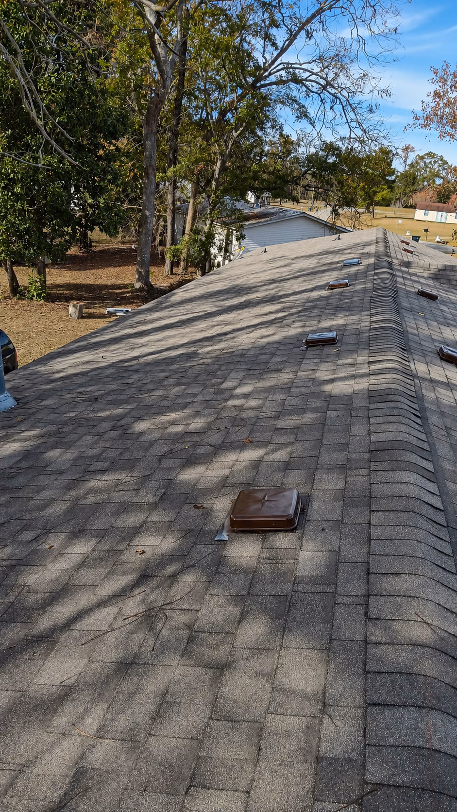 Gray shingled roof with several brown vent covers and trees in the background under a blue sky.