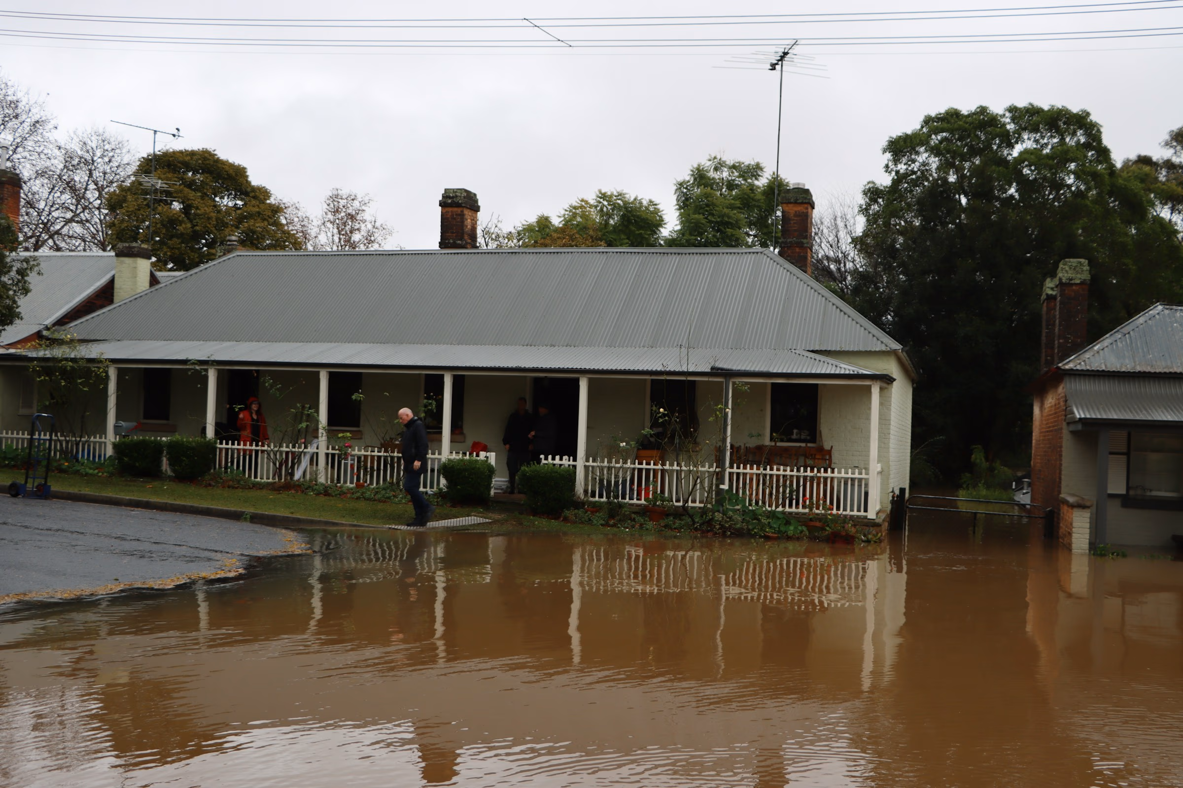 Flooded street with brown water covering road in front of a house with a porch and people standing.